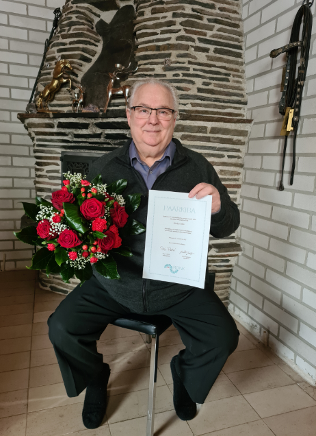 Kuvassa on vuoden 2020 Paarkiba-tunnustuksen saanut Markus Allan. Hän istuu tuolilla ja pitelee käsissään punaista kukkakimppua sekä Paarkiba-kunniakirjaa.